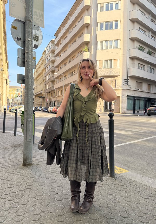 Young woman in green sleeveless top and plaid skirt stands on city sidewalk holding jacket, smiling thoughtfully with finger near lips.