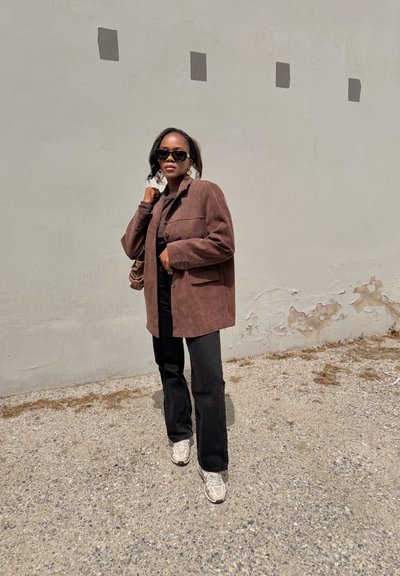 Mujer con blazer marrón de gran tamaño, pantalones negros, zapatillas blancas y gafas de sol, de pie sobre grava frente a una pared beige con detalles cuadrados.