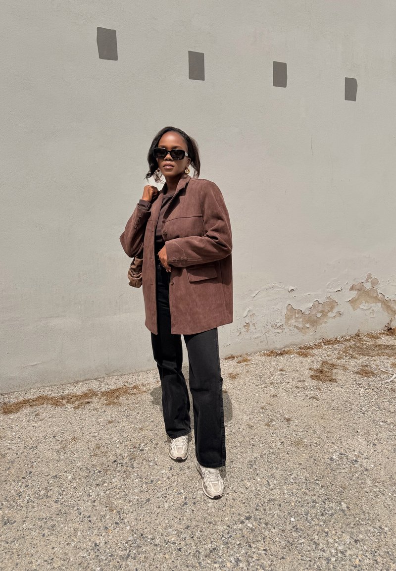 Vrouw draagt een bruine oversized blazer, zwarte broek, witte sneakers en zonnebril, staand op grind tegen een beige muur met vierkante accenten.