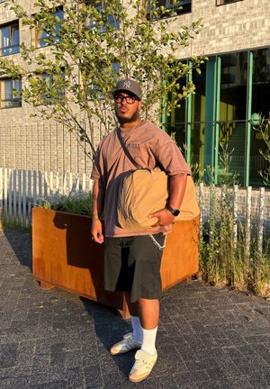 Bolso mensajero marrón con diseño plisado, llevado sobre una camiseta de gran tamaño marrón y pantalones cortos negros. Calcetines blancos con zapatos beige. El fondo presenta vegetación.