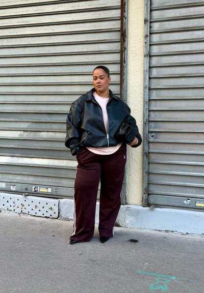 Mujer con chaqueta de cuero negra y pantalones de chándal burdeos de Adidas se encuentra contra persianas de metal cerradas, sosteniendo un bolso de mano negro de mimbre.