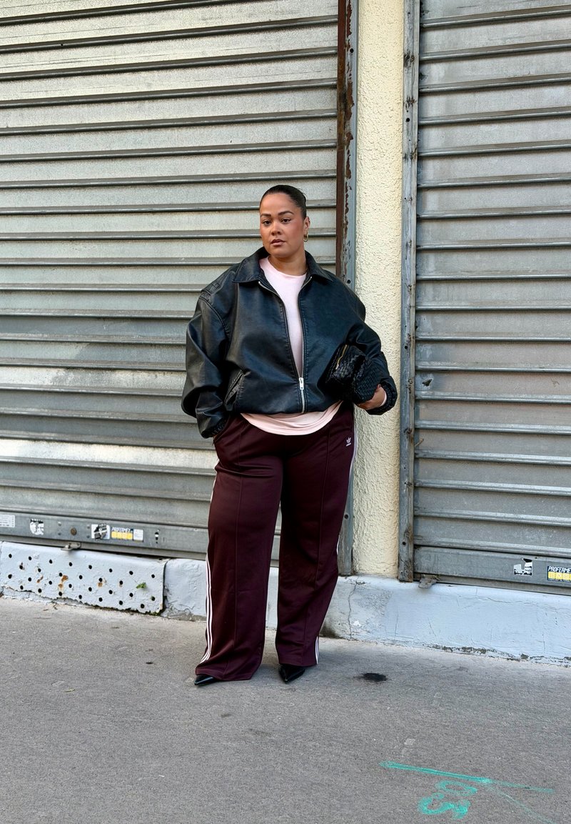 Žena v čiernej koženej bunde a bordových teplákových nohaviciach Adidas stojí proti zatvoreným kovovým roletám a drží čiernu tkanú kabelku.