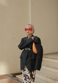 Oversized sort blazer over en sort top, mønstret brun og hvid nederdel, orange håndtaske, røde solbriller og farverigt hår med en blomsteraccessory.