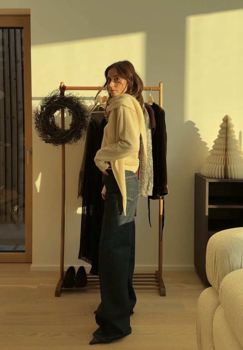 Femme portant un pull jaune et un large pantalon noir se tient près d'un portant en bois avec des vêtements suspendus et une couronne dans une pièce baignée de lumière.