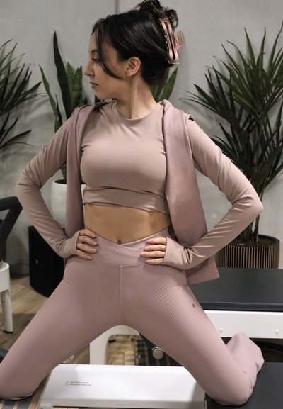 Mujer con ropa deportiva malva arrodillada con las manos en las caderas, mirando hacia la izquierda, en un espacio interior con plantas y equipo de ejercicio.