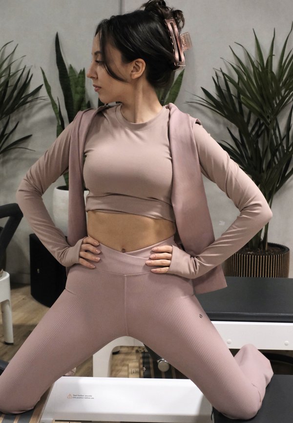 Naissoost, kes kannab lillakast spordirõivastust, on põlvili, käed puusas, vaatab vasakule siseruumis, kus on taimi ja treeningseadmeid.