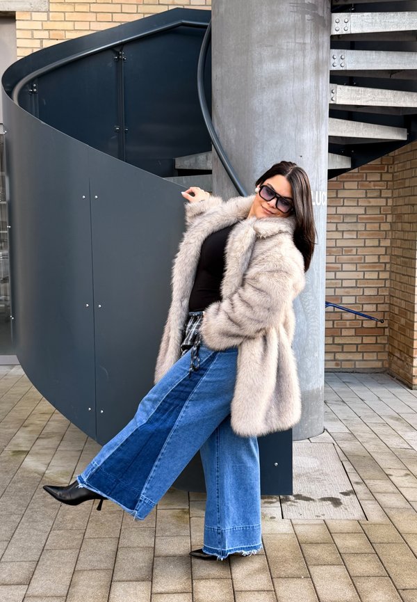 Kvinna som bär solglasögon, en beige pälsrock och vida, tvåfärgade jeans, lutande bakåt vid en spiraltrappa utomhus.