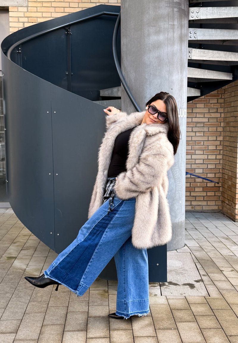 Femme portant des lunettes de soleil, un manteau en fourrure beige et un jean large bicolore, se penchant en arrière près d'un escalier en colimaçon à l'extérieur.