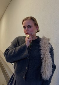 Grauer Wollcardigan mit Knopfverschluss und Kunstfell-Akzenten. Das Model trägt goldenen Schmuck, vor einem strukturierten Hintergrund in neutralen Farben.