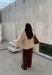 Giacca beige in pelliccia ecologica con maniche larghe, abbinata a pantaloni della tuta color bordeaux con strisce bianche. Sullo sfondo, parete di cemento e scale.