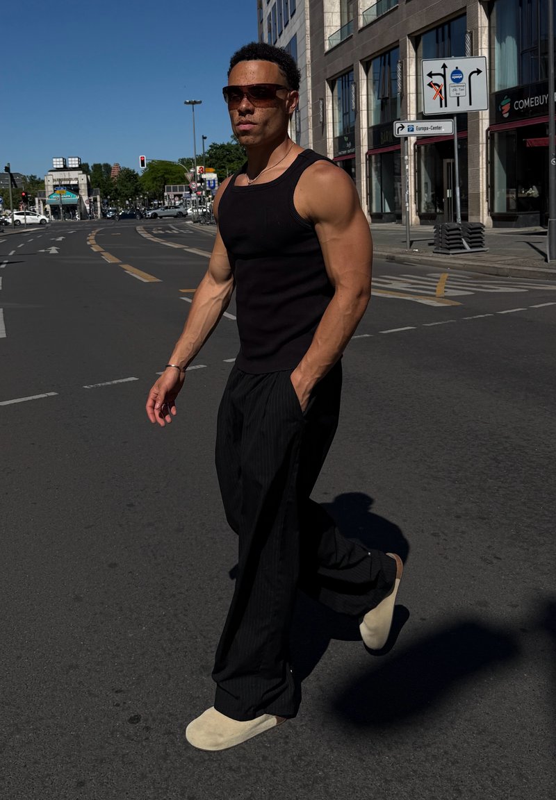 Homme portant des lunettes de soleil, un débardeur noir et un pantalon, traversant une rue en ville avec des bâtiments et des panneaux de signalisation en arrière-plan.