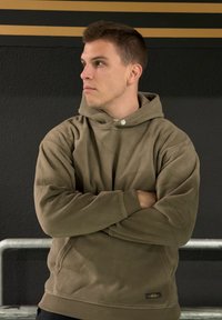 Hanorac din fleece verde măsliniu, cu o croială ușor largă, glugă ajustabilă cu șnur și închidere cu nasturi. Material texturat și manșete striate.