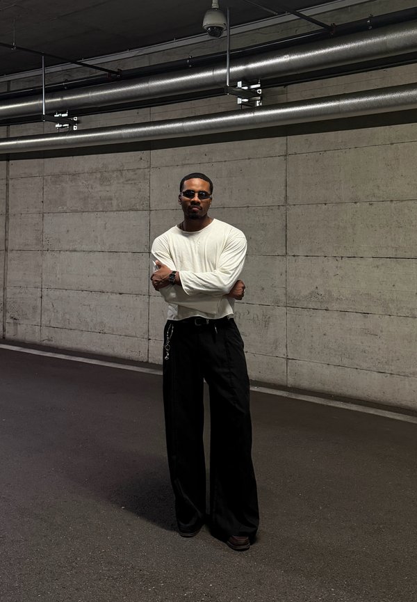 Un homme portant des lunettes de soleil, une chemise blanche à manches longues et un pantalon noir ample se tient les bras croisés dans un passage souterrain en béton.