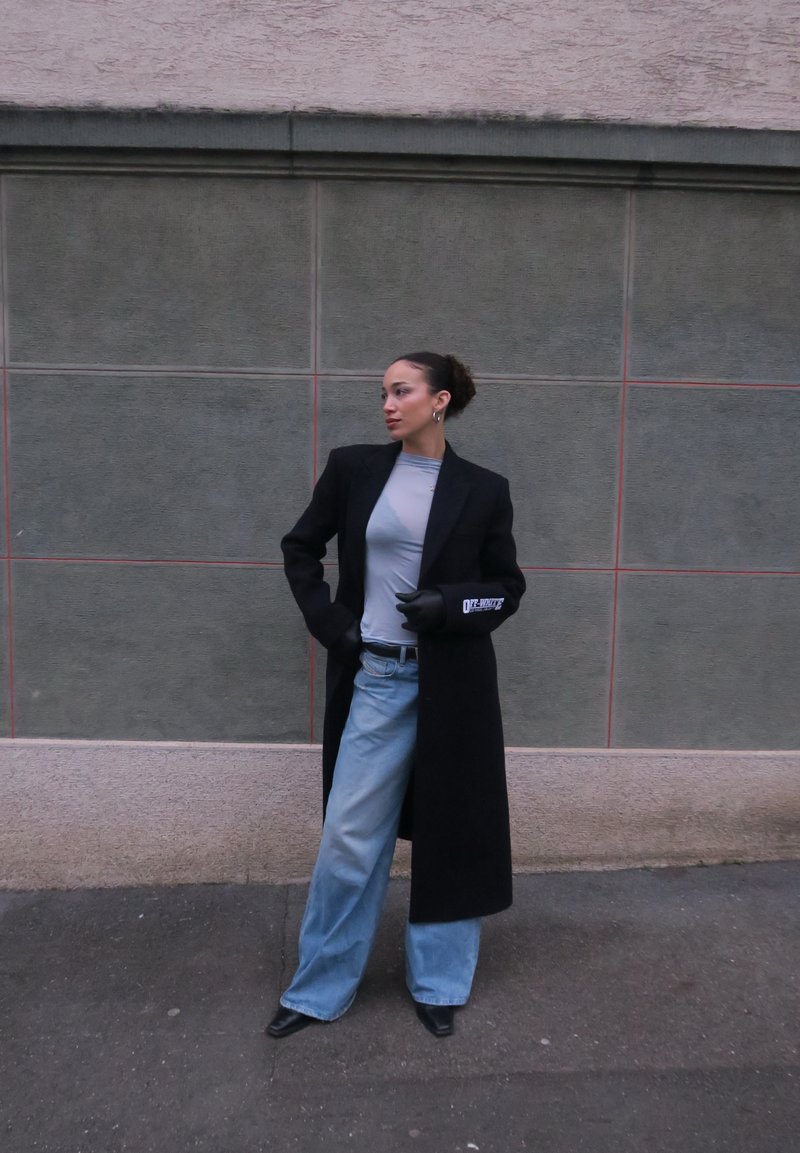 Femme portant un long manteau noir, un haut gris, un jean large bleu, des gants noirs et des chaussures noires à bout pointu, debout contre un mur carrelé gris.