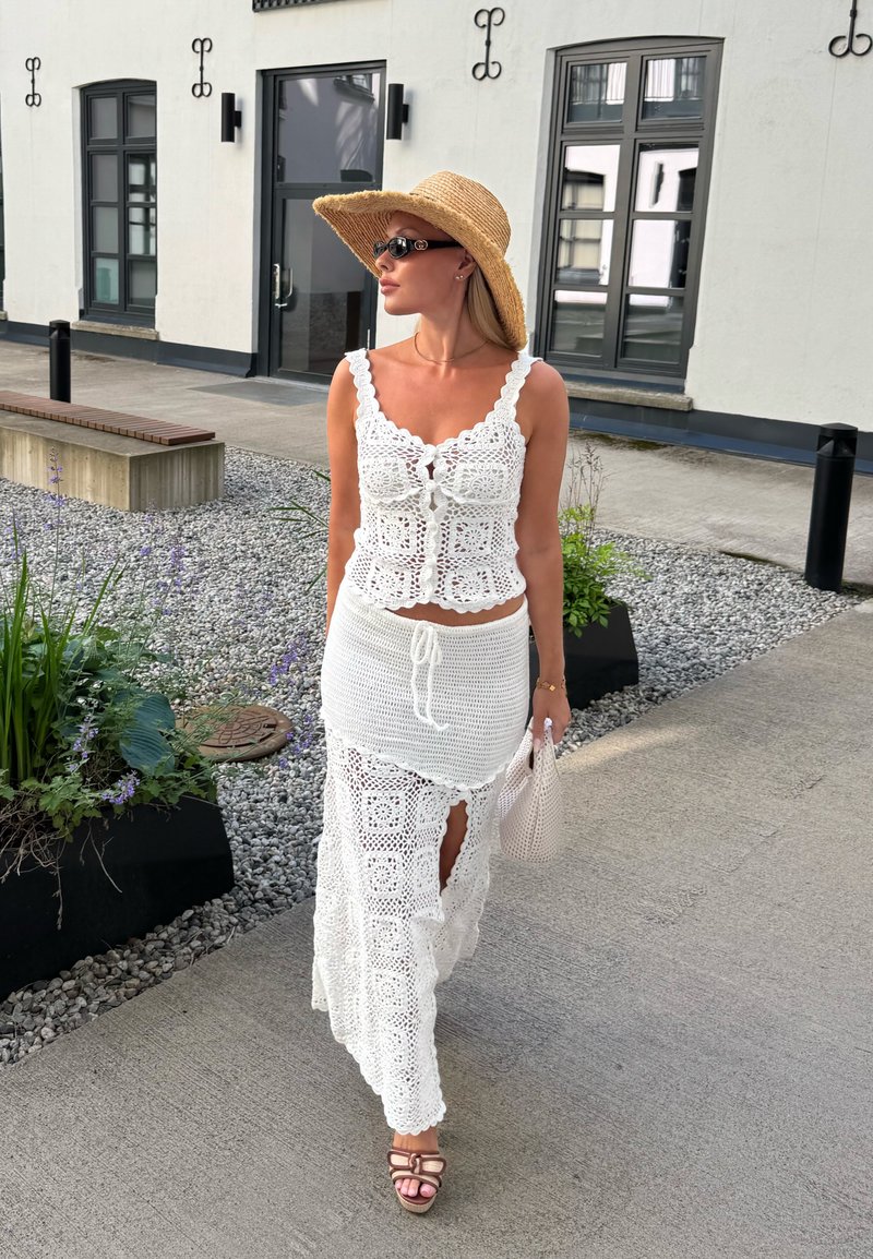 Femme marchant en extérieur portant un haut et une jupe en crochet blanc, un large chapeau de paille, des lunettes de soleil, des sandales et tenant un petit sac.