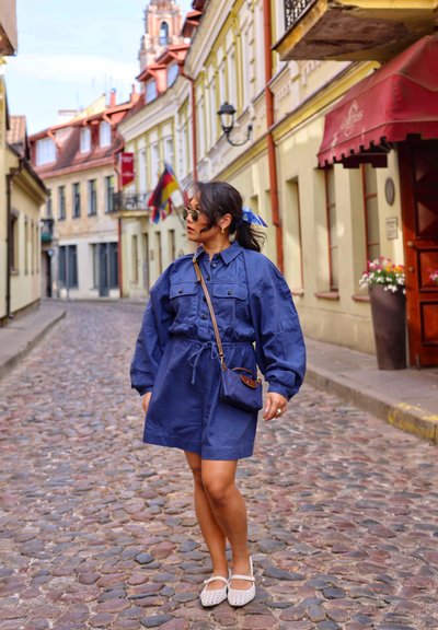 Vestido de algodón azul marino con grandes bolsillos, cintura con cordón y mangas largas. Combinado con zapatos blancos perforados y un pequeño bolso cruzado marrón.