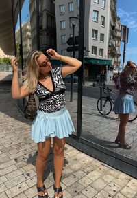 Črno-bela dvobarvna majica s črtami zebre, svetlo modra plisirana krila, črna torbica s nitenjem in črne sandale. Odsevno steklo prikazuje mestno ozadje.