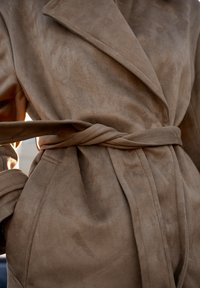 Beige suède trenchcoat met een sjaalkraag, tailleband en zachte textuur, met een losse pasvorm en minimale stikdetails.