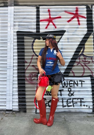 Botas de goma rojas hasta la rodilla combinadas con una camiseta azul tipo polo y una falda negra con estampado de estrellas. Un pequeño bolso rojo con un accesorio verde.