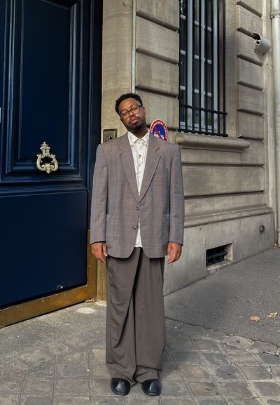 Blazer de cuadros de gran tamaño en marrón y gris, camisa blanca, pantalones grises holgados y zapatos negros. De pie frente a una puerta azul.