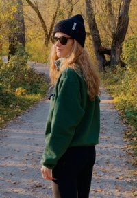 Zielona kurtka polarowa z wysokim kołnierzem, wyposażona w kieszeń na zamek i czarne akcenty, zestawiona z czarnymi legginsami, okularami przeciwsłonecznymi i czapką typu beanie.