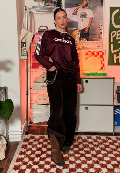 Mujer se encuentra en el interior vistiendo una camiseta de manga larga Adidas color marrón oscuro, pantalones negros con una cadena y botas marrones, con un fondo de habitación decorada.
