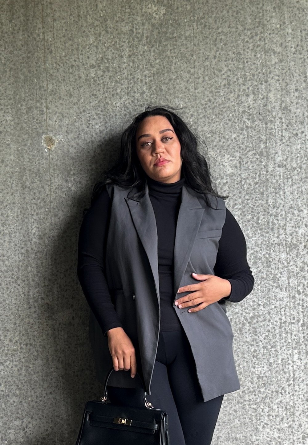 Grå ærmeløs vest over en svart turtleneck, kombinert med svarte leggings. Holder en svart håndveske, satt mot en teksturert grå vegg.