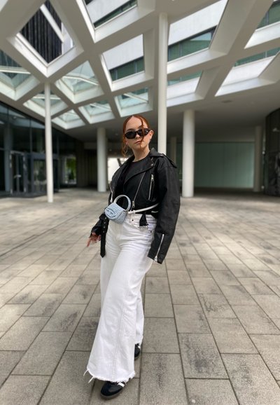 Chaqueta de motociclista de cuero negro, camiseta negra, jeans blancos de pata de elefante y zapatillas negras. Pequeño bolso azul llevado alrededor de la cintura. Fondo urbano.