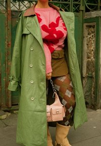 Personne portant un trench-coat vert, un pull rose et rouge, une jupe marron, des collants à losanges, des bottes beige, tenant un sac à main rose devant un portail vert.