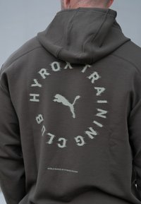 Sweat à capuche vert foncé avec un graphique rond affichant "HYROX TRAINING CLUB" et un logo Puma. Le texte est dans une teinte plus claire près du centre.