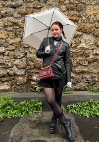 Czarna skórzana kurtka, szary sweter, krótka czarna spódnica, czarne rajstopy, glany. Trzyma białą parasolkę z koronkowym wykończeniem, czerwoną torebkę.