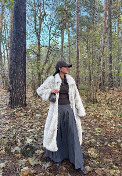 Mujer con un largo abrigo de piel blanco, falda plisada gris, suéter marrón y gorra, de pie en un bosque con hojas caídas y árboles altos.