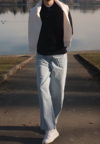 Άτομο που περπατάει σε μονοπάτι δίπλα στη λίμνη φορώντας μαύρο τοπ, ανοιχτό μπλε τζιν, λευκά αθλητικά παπούτσια και ελαφρύ μπουφάν περασμένο στους ώμους.