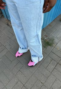 Rozā teksturētas kurpes ar unikālu siluetu, kontrastējot ar gaiši ziliem, brīvi pieguļoši džinsiem, stāv uz bruģētas virsmas.