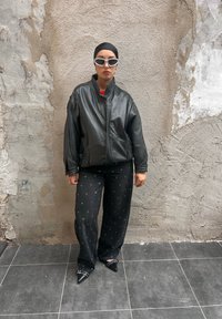 Schwarze Lederjacke mit hohem Kragen, kombiniert mit schwarzen gemusterten Hosen und Spitzenschuhen. Weiße kantige Sonnenbrille rundet den Look ab.