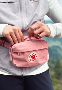 Rosa Fjällräven Kanken belteveske med en glatt tekstur, glidelåslukking og logo-patch. Bært av en person, med justerbar stropp.