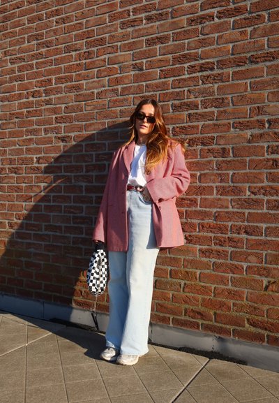 Blazer rosa de gran tamaño, camiseta blanca, vaqueros ajustados de cintura alta en azul claro, bolsa a cuadros en negro y blanco, frente a una pared de ladrillos.
