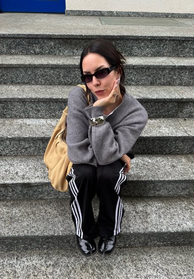 Suéter gris de canalé, pantalones de chándal negros con rayas blancas, zapatos negros y un bolso bandolera beige. Sentado en los escalones de piedra con una puerta azul detrás.