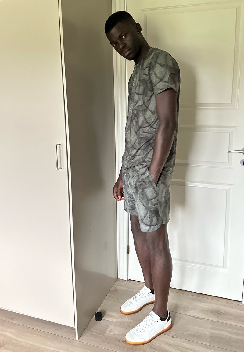 Uomo in piedi all'interno che indossa una camicia grigia a fantasie e pantaloni corti con sneakers bianche, mani nelle tasche, vicino a una porta bianca e a un mobile.