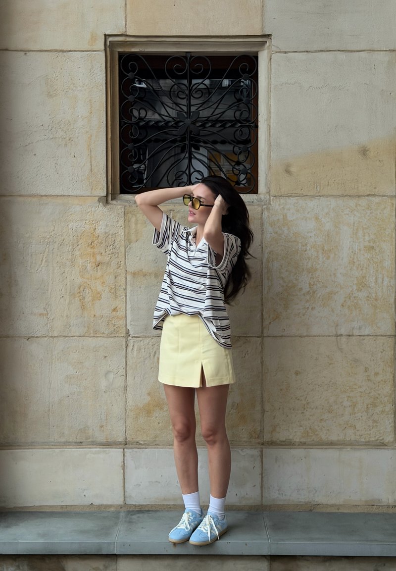 Donna con camicia a righe, gonna gialla e scarpe blu in piedi con le mani tra i capelli davanti a un muro di pietra beige con finestra nera con sbarre.