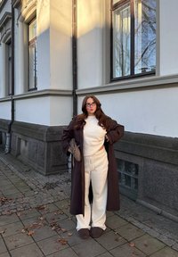 Brun trenchcoat over en kremfarget genser og matchende joggebukser. Brune tøfler, holder en veske; står på en brosteinsbelagt gangvei nær en bygning.