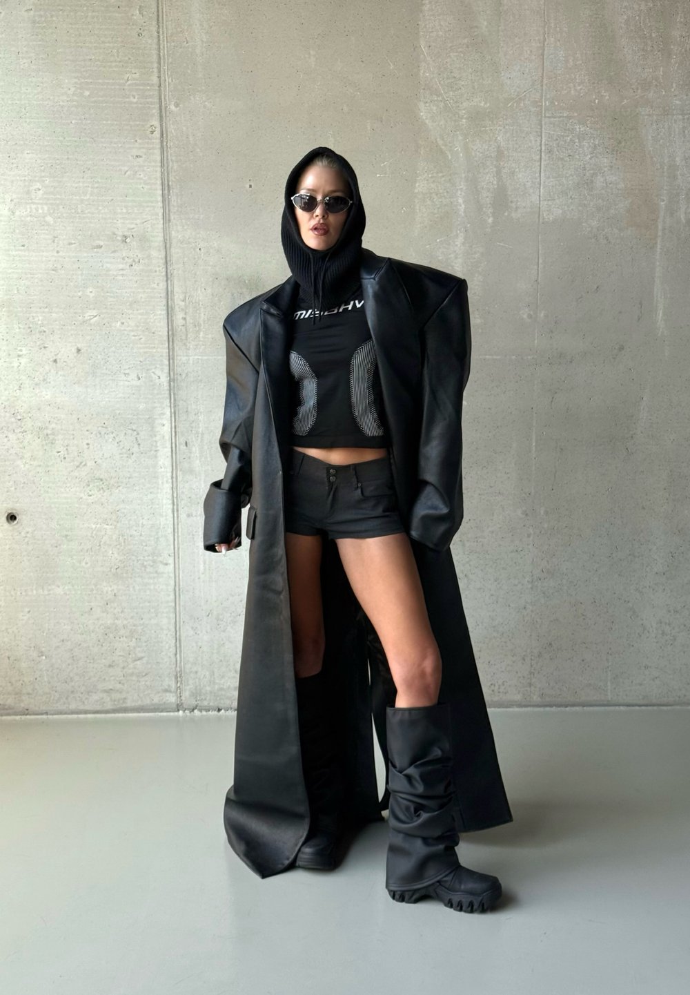 Personne en manteau en cuir noir surdimensionné, top court noir, short, capuche, lunettes de soleil et bottes noires décontractées devant un mur en béton.