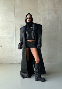 Personne en manteau en cuir noir surdimensionné, top court noir, short, capuche, lunettes de soleil et bottes noires décontractées devant un mur en béton.