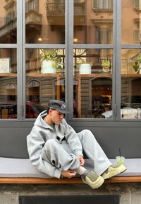 Szara, oversize'owa bluza z kapturem i dresy, zielone buty z czarnymi akcentami, siedząca na szarej ławce, zielony napój obok, nowoczesne tło kawiarni.