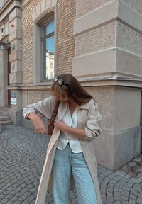 Μπεζ ελαφρύ παλτό τύπου trench πάνω από λευκό μπλουζάκι, συνδυασμένο με ανοιχτό μπλε παντελόνι σε ευρεία γραμμή. Τα αξεσουάρ περιλαμβάνουν σκούρα γυαλιά ηλίου και καφέ τσάντα.