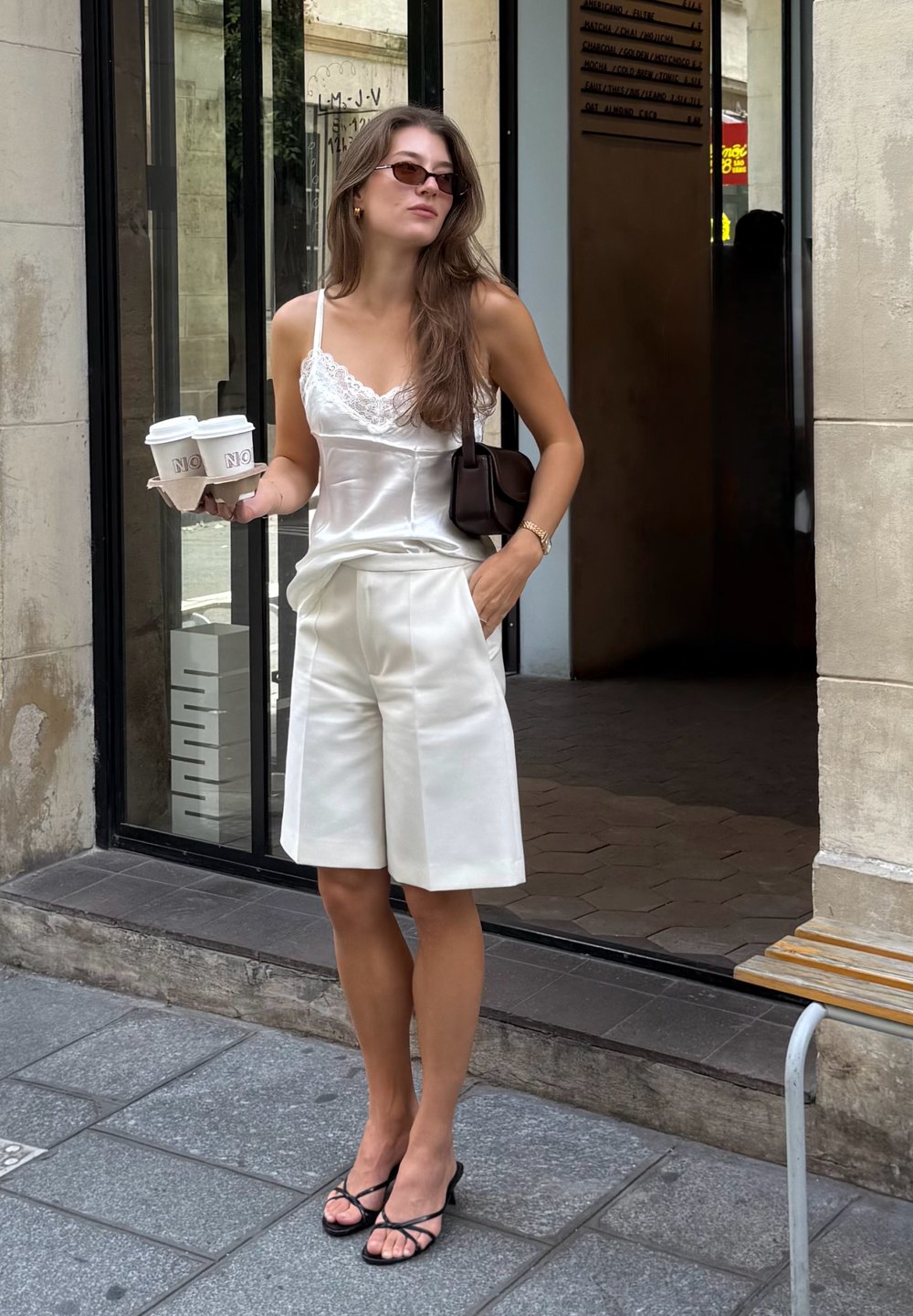 Bijela satenasta majica na bretele s čipkastim rubom, bijele krojene kratke hlače, crna torbica, crne sandale s trakama, drži dva šalice kave u kartonskom nosaču.