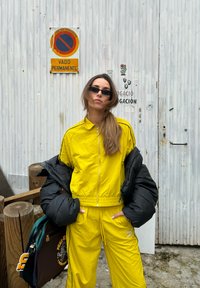 Κίτρινο φόρμα με ανοιγόμενο μπουφάν, συνδυασμένο με μαύρο μπουφάν με επένδυση. Το μοντέλο κρατάει μια καφέ και πράσινη τσάντα. Το φόντο περιλαμβάνει ένα ξύλινο στύλο.