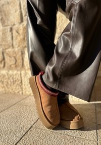Chaussures à enfiler en daim marron avec bordure tricotée rouge et blanche, portées avec un pantalon en cuir marron foncé, debout sur un sol en pierre texturé.