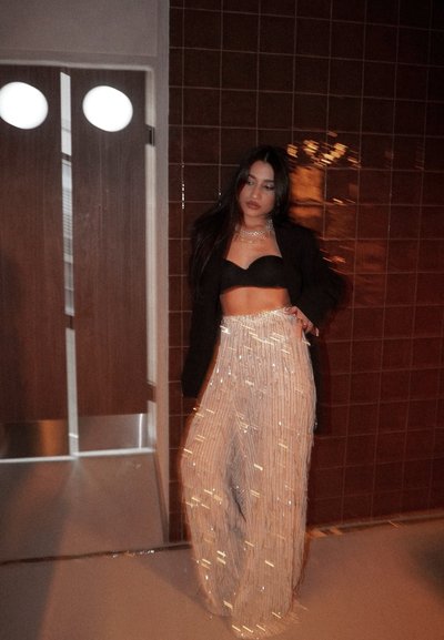 Mujer con crop top negro, chaqueta negra y pantalones anchos brillantes, de pie contra una pared marrón con azulejos cerca de unas puertas dobles de madera.