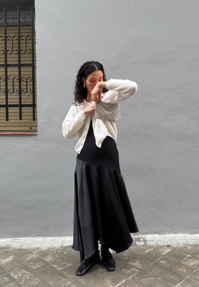 Cárdigan de punto blanco sobre un vestido negro sin mangas con dobladillo acampanado. El conjunto incluye zapatos negros y un bolso plateado texturizado.
